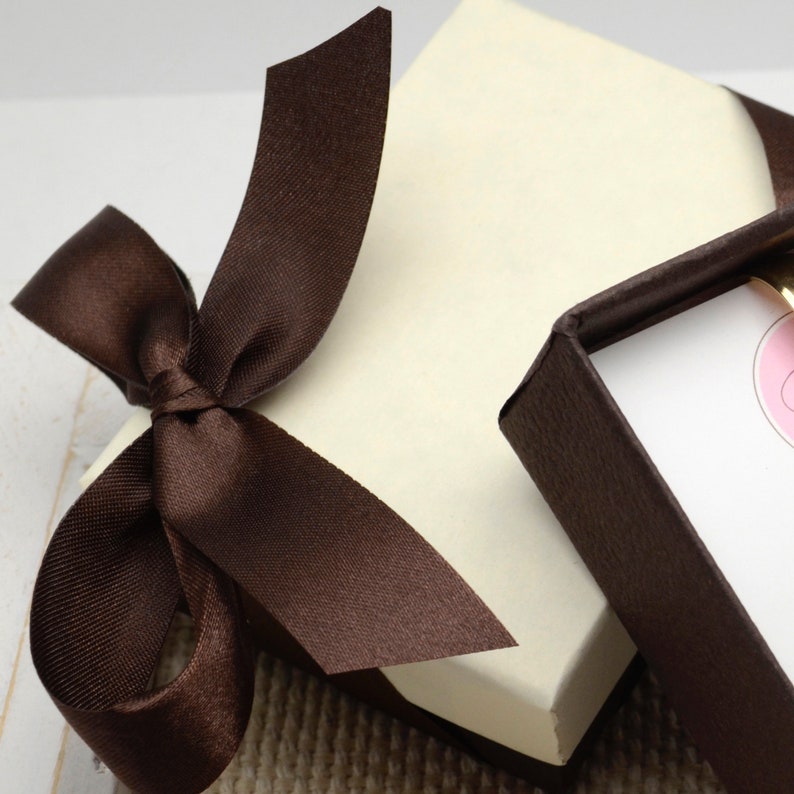May include: A cream-colored gift box tied with a dark brown satin ribbon and bow. The box is partially open, revealing a glimpse of the contents. The overall aesthetic suggests a presentation for a special occasion.