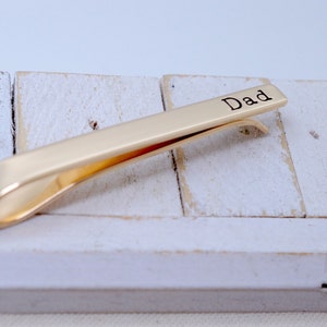 May include: A gold-colored tie clip with the word "Dad" engraved on the top. The clip is resting on a white, weathered wooden surface. The tie clip is a simple, elegant accessory.