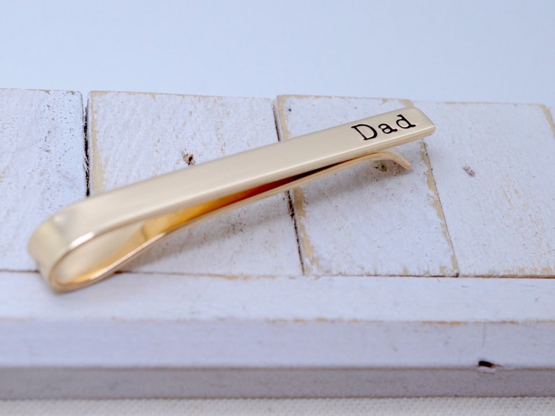 May include: A gold-colored tie clip with the word "Dad" engraved on the top. The clip is resting on a white, weathered wooden surface. The tie clip is a simple, elegant accessory.