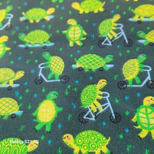 May include: Dark teal fabric with a repeating pattern of yellow-green turtles on skateboards, scooters, and bicycles. The turtles have patterned shells and are set against a background of small blue and green shapes.