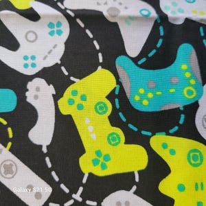 May include: A black fabric with a repeating pattern of colorful video game controllers. The controllers are in shades of green, yellow, and white. The fabric has a stitched design around each controller.