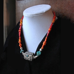 May include: A colorful necklace with orange, red, and turquoise beads. The necklace features a silver-colored, square pendant with an embossed design. The necklace is displayed on a white mannequin head against a black background.