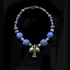 May include: A necklace featuring a series of blue and patterned beads, interspersed with small metallic spheres. The centrepiece consists of two silver-toned bell-shaped charms hanging above a small, triangular, metallic pendant with a hook clasp.