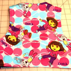 May include: A piece of fabric featuring a repeating pattern of pink and blue polka dots with cartoon characters. The characters include a girl and a monkey, with the word "DORA" printed on the fabric.