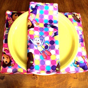 May include: A yellow plate rests on a colorful placemat and is topped with a matching napkin. The placemat and napkin feature a repeating pattern of cartoon characters and polka dots in pink, purple, yellow, and blue.
