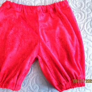 May include: Bright red, elastic-waist bloomers. The fabric has a soft, velvety texture. The legs are gathered at the cuffs. The garment is likely for a baby or toddler. The background is a white, quilted surface.