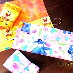 May include: A collection of colorful fabric pouches and cases. One pouch features SpongeBob SquarePants with the text "It's All Good". Another has Eeyore from Winnie the Pooh. The items are in shades of yellow, orange, pink, and blue.