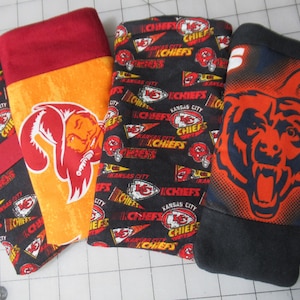 May include: Four fabric items with sports team logos. One is orange with a red logo, another is black with red and white logos, and the last is black with a blue and orange bear logo. The fourth item is black with red and white logos.