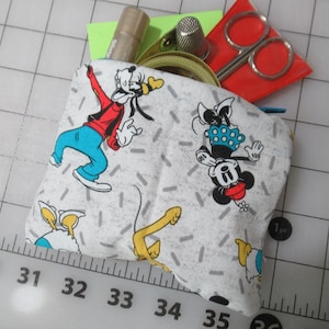 Peut inclure: Une pochette en tissu blanc avec un motif à pois gris et blanc. La pochette présente des images de personnages de dessins animés de Mickey Mouse et de Dingo. La pochette est ouverte et montre une variété d'accessoires de couture, y compris des ciseaux, du fil et un dé à coudre.