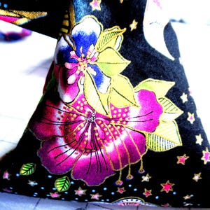 May include: Close-up of a black fabric item with a vibrant floral pattern. The design features large pink and blue flowers with gold accents, set against a backdrop of stars and dots. The item appears to be a decorative element.