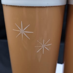 May include: Set of vintage-style drinking glasses. The glasses are a light brown color with a white rim and starburst designs. The glasses are cylindrical and appear to be made of glass or a similar material.