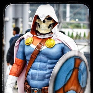 Puede incluir: Un personaje disfrazado que lleva una capa blanca con capucha y una máscara de calavera, un traje azul y blanco y un cinturón de cuero marrón. El personaje sostiene un escudo azul y naranja con un borde plateado.