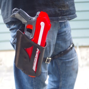 Könnte beinhalten: Ein schwarzes Lederholster mit einer roten und weißen Spielzeugpistole, die an einem Gürtel befestigt ist. Das Holster wird an der rechten Hüfte einer Person getragen, die eine blaue Jeans trägt.