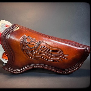 Op de afbeelding: Een bruine leren holster met een paardenkopontwerp. Een wit en rood speelgoedpistool zit in de holster.