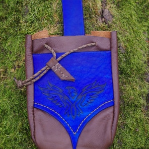 May include: A brown leather pouch with a blue leather flap featuring an eagle design. The pouch has a drawstring closure and a blue leather strap.
