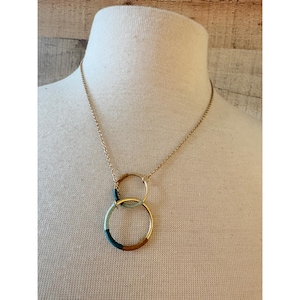 May include: A gold-toned chain necklace featuring two interlocking circular pendants. The smaller top ring is plain gold, while the larger bottom ring is accented with segments of teal, light blue, and brown enamel, creating a layered geometric design.