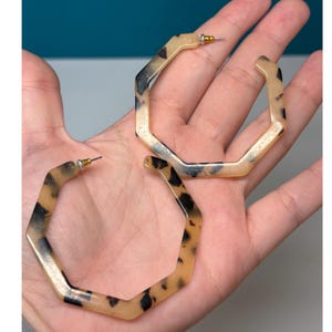 May include: A pair of hexagonal hoop earrings featuring a translucent beige and brown tortoiseshell pattern. The earrings are constructed from a smooth resin material and include small metallic posts with silver and gold-toned hardware.