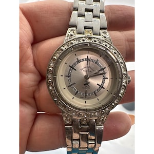May include: A silver-toned Beverly Hills Polo Club quartz watch with a round face and a crystal-studded bezel. The watch features a silver dial with black hands and hour markers. The watch band is also silver-toned.
