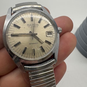 May include: A vintage Baron automatic wristwatch with a silver-tone case and a light gold-colored face. The watch face displays the brand name, "Baron 17 Jewels", and the word "Automatic". The watch has a date display at the 3 o'clock position and a silver-tone expanding band.