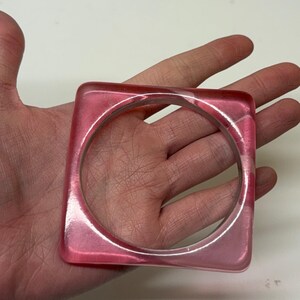 May include: A square-shaped bangle bracelet made of translucent pink resin. The accessory features a smooth, polished finish with a circular inner opening and a distinct square outer perimeter, showcasing a consistent light pink hue throughout the material.