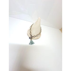 May include: A bracelet featuring a metallic chain with circular links, a toggle clasp, and a light blue tassel charm. The chain is constructed from a dark, antique-toned metal, and the tassel is attached to the clasp with a small, dark bead.