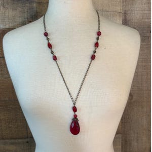 May include: A long necklace featuring a bronze-toned chain with interspersed red beads and small metallic spacers. The necklace terminates in a central pendant consisting of a small red bead topped by a larger, faceted, teardrop-shaped red stone.