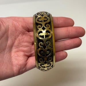 May include: A wide cuff bracelet made of polished gold-tone metal featuring an intricate, openwork filigree pattern with symmetrical scroll and heart-like motifs. The design consists of a repeating cutout structure that reveals a dark interior.