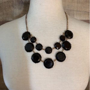 May include: A multi-layered statement necklace featuring round, faceted black stones set in gold-toned metal. The design consists of two tiers of varying sized circular black gems connected by delicate gold-toned chain links, creating a layered look.
