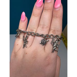 May include: A silver-toned metal charm bracelet featuring four distinct dangling charms. The charms include two winged cherub figures, a heart shape with an arrow, and a small star-shaped charm, all attached to a classic oval link chain.