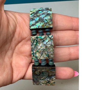 May include: A bracelet featuring three square tiles inlaid with iridescent abalone shell in shades of green, blue, and brown. The tiles are connected by small, dark, cylindrical beads arranged in rows of three between each square segment.