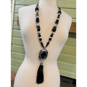 May include: A long black and white beaded necklace with a large oval pendant and a black tassel. The necklace features a variety of beads, including black oval beads, white and black patterned beads, and small silver-colored beads.