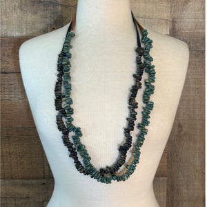 May include: A multi-strand necklace featuring small, irregular, tumbled beads in shades of dark brown and teal green. The beads are strung on thin, dark leather cords that connect to tan leather accents at the top of the necklace structure.
