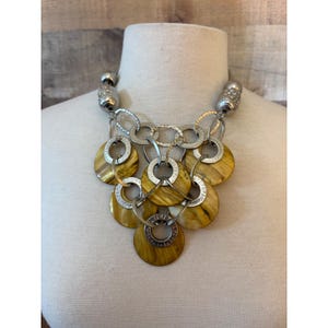 May include: A statement necklace featuring multiple silver-toned metal rings and circular, amber-colored shell discs. The design consists of layered, interlocking metallic links and polished, warm-toned circular pendants arranged in a cascading pattern.
