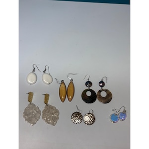 May include: A collection of six pairs of dangle earrings featuring various designs, including smooth white oval drops, amber-colored marquise shapes, dark iridescent circular discs, textured gold-tone metal rounds, clear faceted shapes, and small blue iridescent circles.