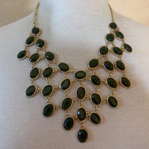 May include: A gold-tone statement necklace featuring a cascading bib design composed of multiple dark green oval-shaped cabochon stones set in metal frames, connected by delicate gold-tone links that form a structured, web-like pattern.