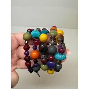 May include: A multi-strand wrap bracelet featuring an assortment of colorful beads in shades of yellow, orange, turquoise, olive green, dark red, purple, and grey. The design includes smooth round beads, ribbed textured beads, and metallic accents.