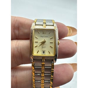 May include: Oleg Cassini quartz watch with a rectangular gold-tone and silver-tone case and band. The watch face is cream-colored with gold-tone hands and markers. The date is displayed at the bottom. The watch is a classic accessory.