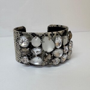 May include: A wide cuff bracelet featuring a snakeskin pattern in shades of gray, black, and white. The surface is embellished with an array of faceted clear, white, and smoky gray crystals in various shapes, including round and teardrop cuts.