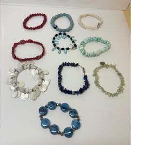 May include: A collection of ten assorted bracelets featuring various materials including red beads, blue stone beads with an evil eye charm, white shell fragments, red oval beads, blue and black accents, light green shell chips, white shell discs, blue lapis lazuli chips, pale green stone chips, and large blue glass beads.
