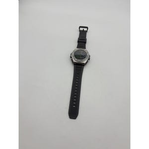 May include: A black digital watch with a silver-colored bezel and a black rubber strap. The watch face displays a digital time readout. The watch is lying on a white surface.