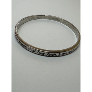 May include: A silver-toned metal bangle bracelet with a smooth, polished finish. The outer surface of the band features engraved black lettering that reads, "three things that last: faith, hope, and love." The bracelet has a simple, circular design.