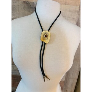 May include: A bolo tie featuring a black braided cord with metallic tips. The central slide is a polished, irregular tan stone piece accented with a small, circular gold-toned setting containing a deep red stone surrounded by tiny clear crystals.