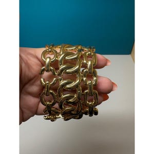 May include: A gold-toned bracelet featuring three rows of interconnected polished metal links. The design consists of a central row of thick, rounded oval links flanked by two outer rows of smaller, rectangular-shaped chain links in a bright finish.