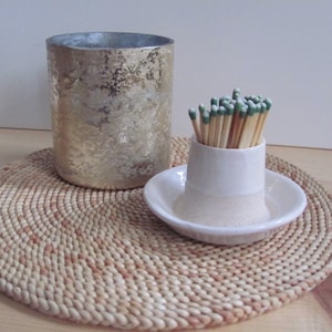 May include: A white ceramic matchstick holder with a round base and a cylindrical top, filled with wooden matches with green tips. The holder is sitting on a woven straw placemat next to a silver metallic candle holder.