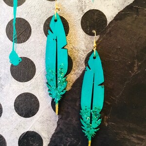 Puede incluir: Un par de pendientes de plumas de color azul turquesa con detalles de purpurina dorada y ganchos dorados.