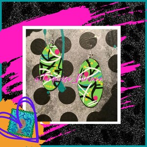 Pode incluir: Um par de brincos ovais com um design abstrato verde, preto, branco, rosa e amarelo. Os brincos estão sobre um fundo de bolinhas pretas e brancas.