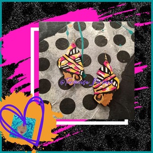 Puede incluir: Un par de pendientes coloridos pintados a mano que representan el rostro de una mujer con un turbante estampado en amarillo y rosa. Los pendientes están sobre un fondo de lunares negros y blancos.