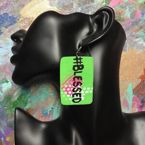 Puede incluir: Pendiente de madera verde con el texto "#BLESSED" en letras negras. El pendiente tiene un diseño abstracto rosa y blanco.
