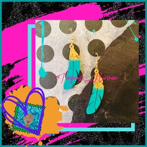 Puede incluir: Un par de pendientes de plumas turquesa y dorado. Las plumas están pintadas con un acento dorado y tienen una base turquesa. Los pendientes cuelgan de ganchos plateados.