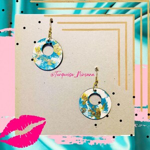 Puede incluir: Un par de pendientes redondos con un agujero central, pintados en blanco, azul y dorado. Los pendientes cuelgan de ganchos dorados. El texto "Turquoise Nirvana" es visible.